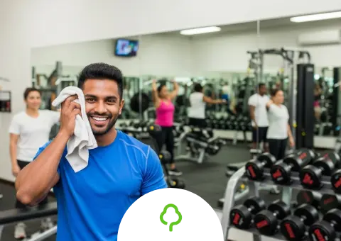 Fitness gym owner Izak Reddy smiling and wiping his face with a towel after a workout, with other members exercising on various machines in the background of the busy gym.
