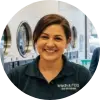 Laundromat owner Lindi Fourie smiles warmly at the camera while folding a stack of fresh blue towels in her clean and busy laundromat, with customers using the washing machines in the background.